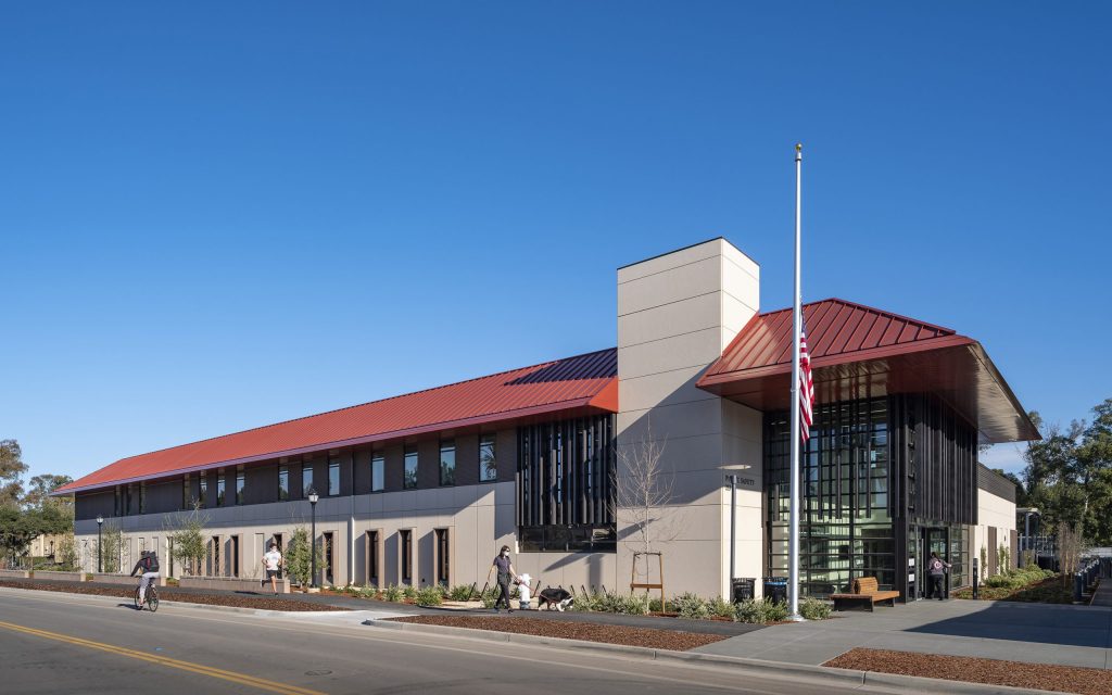 The two-story building, adorned with a red gable roof and large glass windows, showcases the craftsmanship of California-based general contractor Level 10 Construction. An American flag waves proudly out front as people walk and cycle by, framed by trees and clear blue skies.