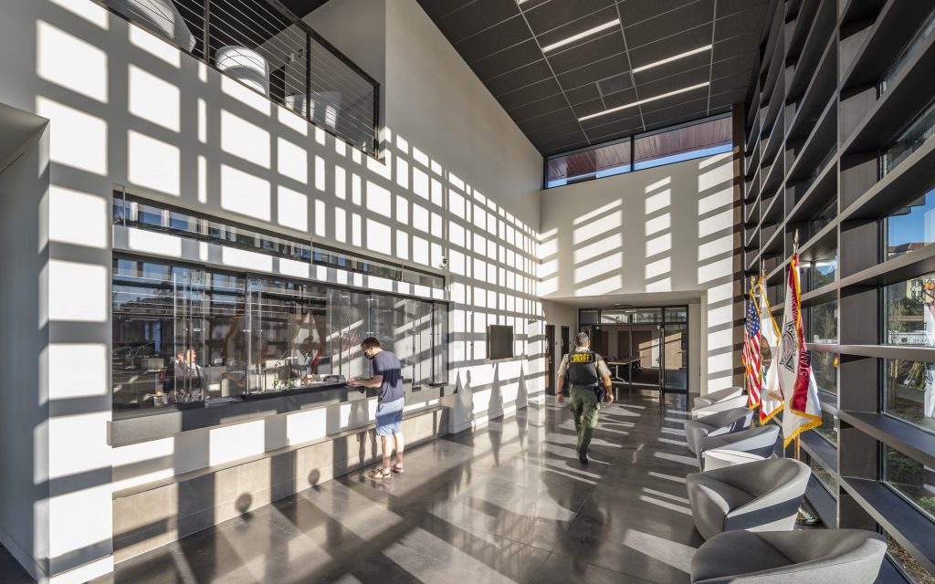 A sunlit lobby with patterned shadows on the walls, designed by Level 10 Construction, shows two people at a counter with large glass windows. American and state flags stand in the corner. Modern chairs line the right side under a high ceiling in this California-based general contractor's masterpiece.