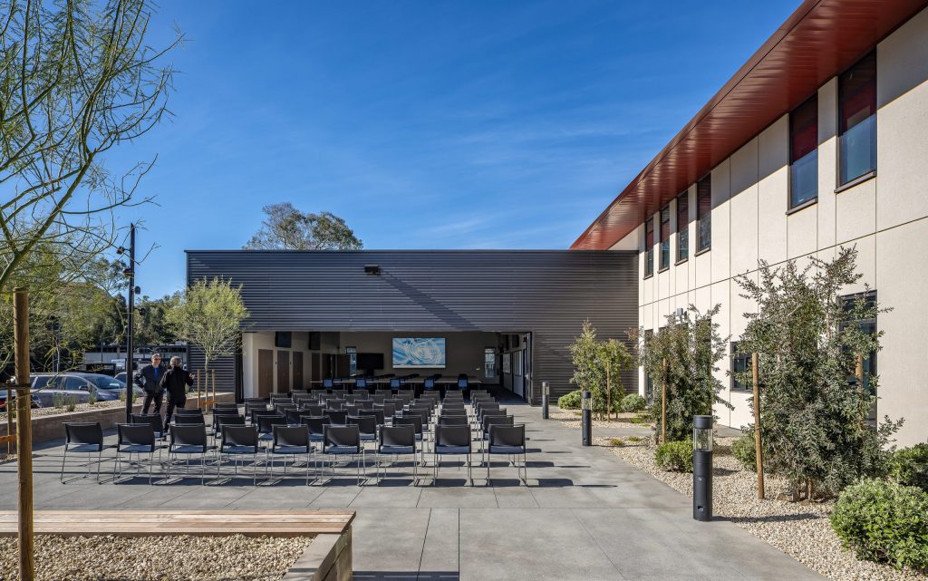 Outdoor gathering space with rows of empty chairs facing a modern building featuring a large screen. The sunny setting, beautifully designed by a California-based general contractor, is enhanced by trees and shrubs lining the concrete area. Two people stand nearby under the clear blue sky.