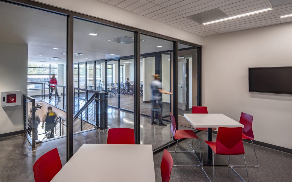 A modern office area, crafted by California-based general contractor Level 10 Construction, features glass walls and square white tables surrounded by red chairs. People walk along a hallway and descend stairs. A wall-mounted TV is visible on the right, and the polished concrete floor gleams below.