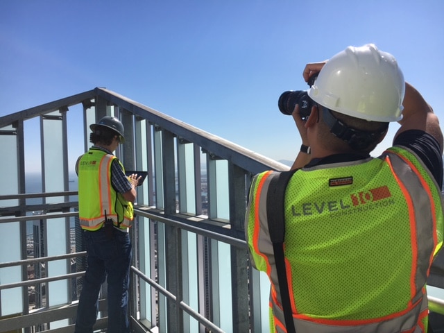 Two construction workers wearing safety vests and helmets are on a high-rise rooftop. One is taking photos with a camera, and the other uses a tablet, both overlooking the cityscape. They are part of Level 10 Construction, a California-based general contractor known for its precision and innovation.