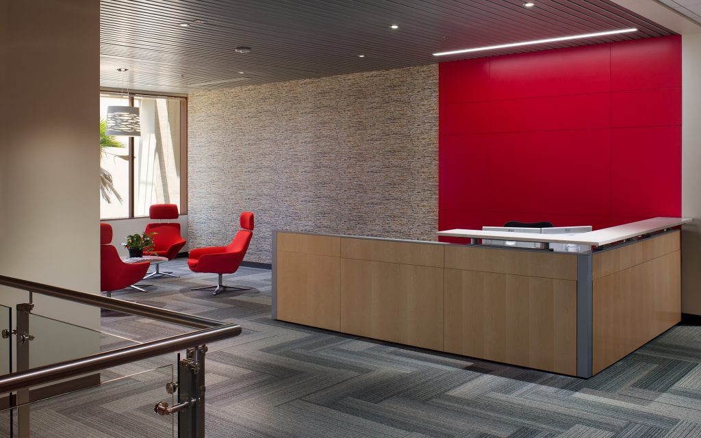 The modern office reception area, designed by California-based general contractor Level 10 Construction, features a wooden desk, red accent wall, and two red chairs with a small green plant on a table. A large window floods the space with natural light over the patterned carpet.