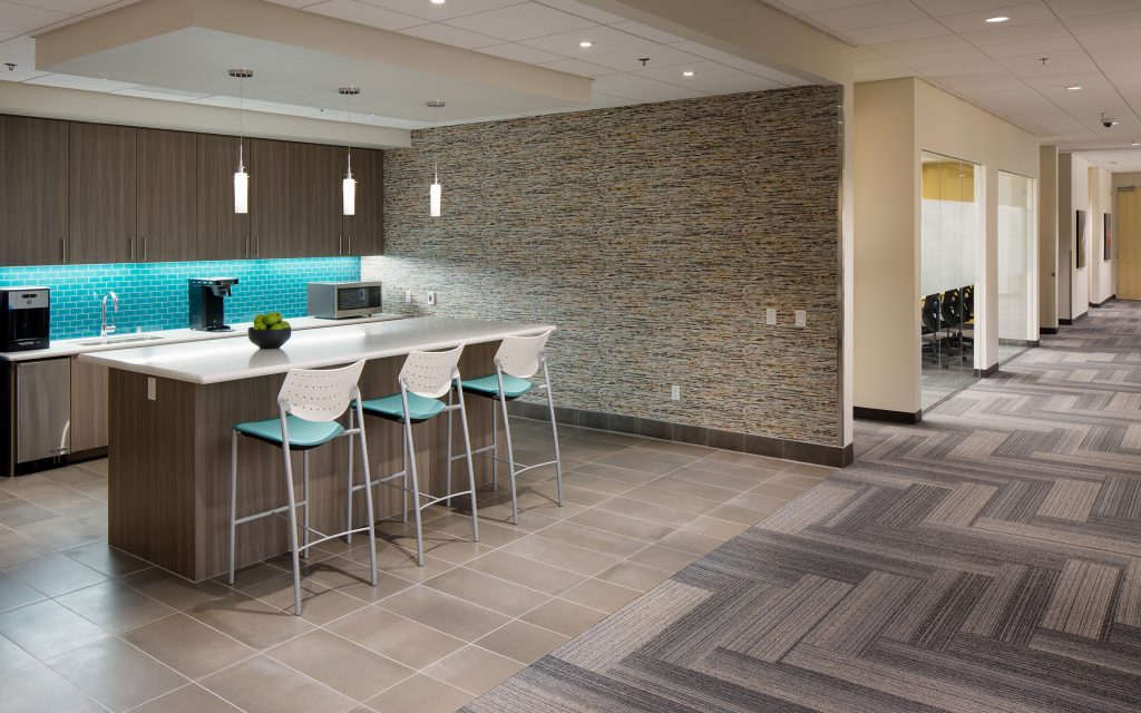 The modern office kitchenette, crafted by Level 10 Construction, features a counter with three stools and overhead lights. A textured wall and blue tile backsplash complement the coffee machine and microwave. An adjoining hallway with carpet tiles leads to a glass-walled meeting room.