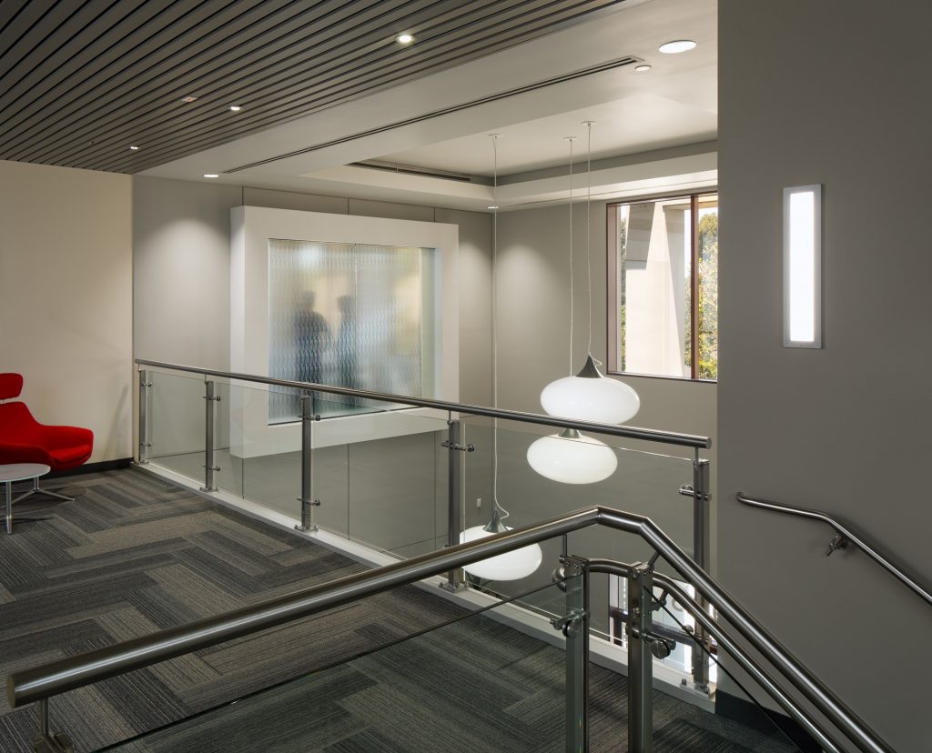 A modern office interior designed by Level 10 Construction features glass railings and a gray carpeted floor. Three white pendant lights hang from the ceiling. A blurred figure is visible through a frosted glass wall near a red chair, as natural light streams in from a window.