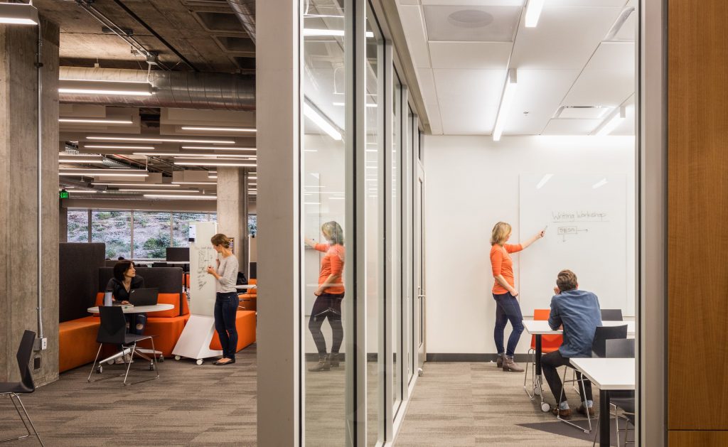 A modern office space with glass walls, crafted by California-based general contractor Level 10 Construction. On the right, two people collaborate at a whiteboard, while on the left, three individuals engage in a discussion around small tables with laptops. The room is well-lit with overhead lights.