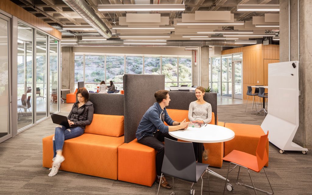 Modern office space with people working. One person is on an orange couch using a laptop, while two others sit at a round table having a discussion. Designed by Level 10 Construction, the room features glass walls, exposed ceilings, and a whiteboard.
