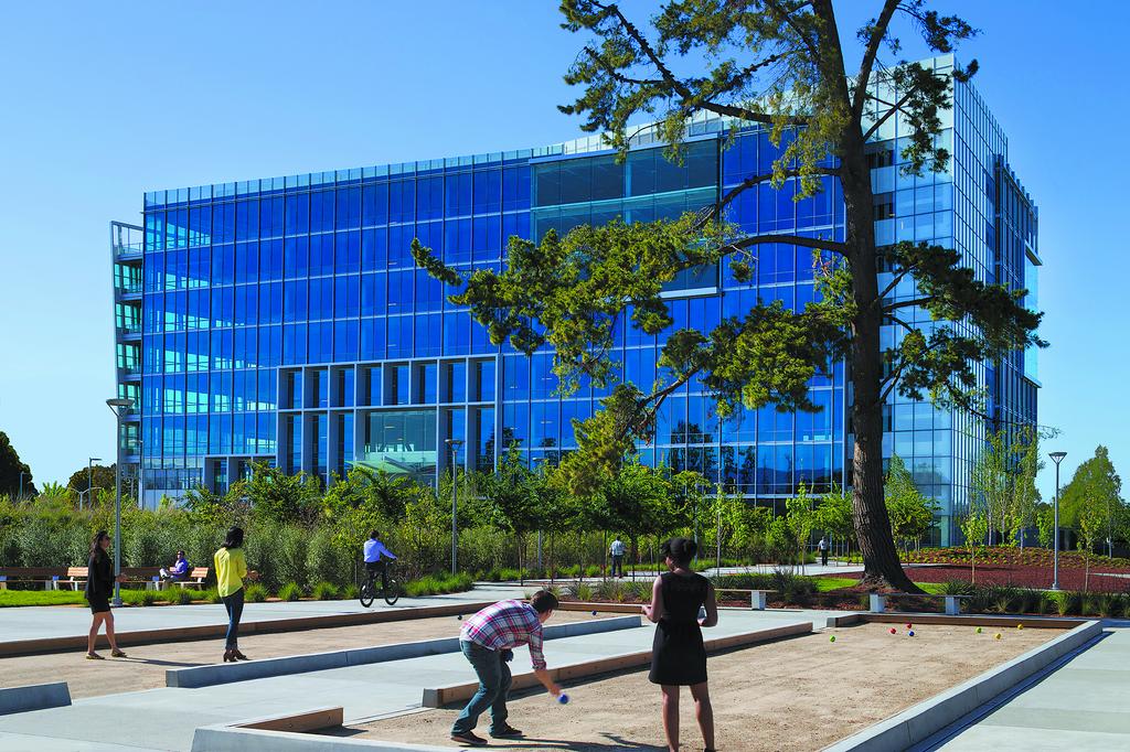Bocce ball enthusiasts enjoy a landscaped area in front of a modern glass office building constructed by Level 10 Construction, with a cyclist passing by in the background. Tall trees and clear blue skies frame this California scene perfectly.