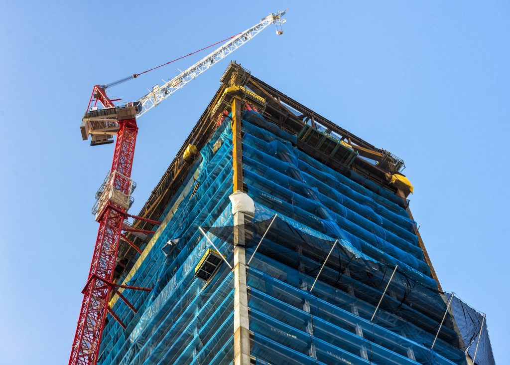 A tall building under construction, led by California based general contractor Level 10 Construction, is covered with blue protective netting. A red and white crane stands next to the structure against a clear blue sky.