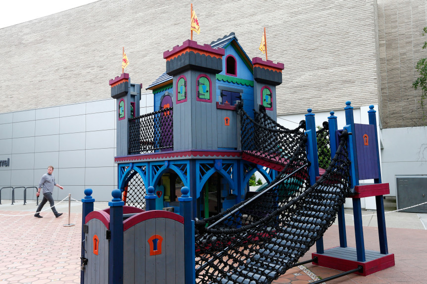 A colorful playground castle, assembled by a California-based general contractor, features towers with red roofs, blue supports, and rope bridges. A person walks nearby in a paved area, and the background shows gray and beige buildings.