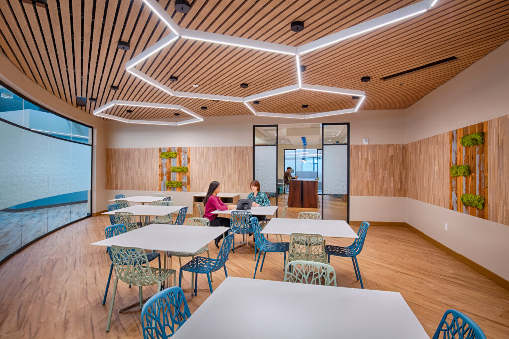 collaborative café space with wood slat ceiling and geometric LED lighting, light wood flooring, modern tables with blue and green chairs, living moss wall accents