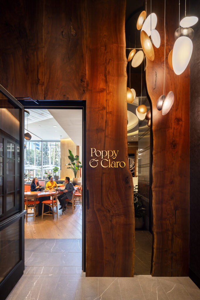 Entrance to Poppy & Claro restaurant featuring live-edge wood wall panels, custom pendant lighting, illuminated signage, and views into the contemporary dining space.
