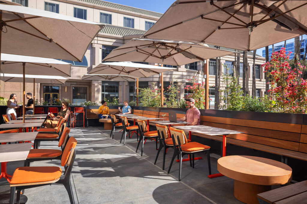 Outdoor patio at Poppy & Claro restaurant featuring contemporary dining tables, shaded umbrella seating, landscaped planters, and a welcoming hospitality environment.