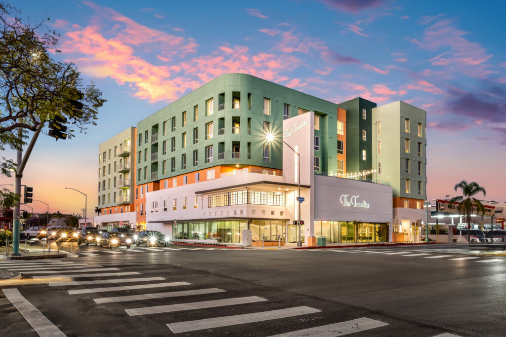Exterior view of The Teralta, a modern multi-story apartment building on a city corner at sunset, featuring colorful фасade, ground-floor retail space, and street activity with cars and pedestrians.