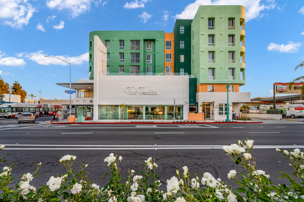 Street-level view of The Teralta, a contemporary multi-story apartment building with a green and orange fасade, ground-floor retail, and landscaped frontage along a city street.