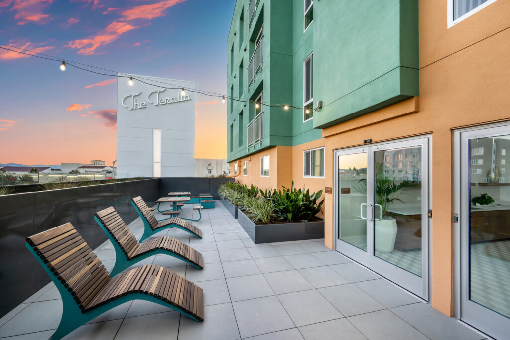 Rooftop terrace at a modern apartment community featuring lounge seating, planters, string lights, and views at sunset, with The Teralta building signage visible.