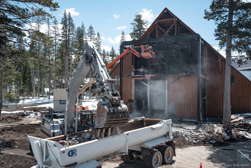 Courtesy of Sugar Bowl Resort An excavator demolishes part of a large timber building at a mountain resort while workers on an elevated lift cut through the steel framework. Dust rises from the demolition as debris is loaded into a dump truck. Snow patches, tall pine trees, and a clear blue sky surround the active construction site.