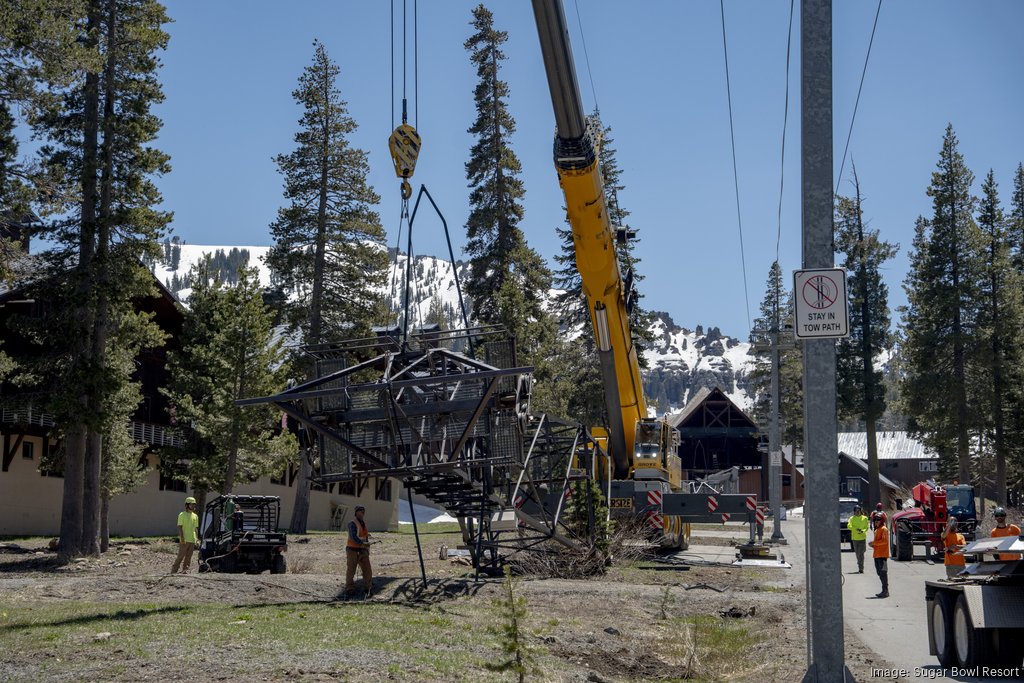 Courtesy of Sugar Bowl Resort A construction crew uses a large crane to lift and position a steel ski lift tower structure at a mountain resort. Workers guide the suspended framework from the ground while snow-covered slopes, pine trees, and lodge buildings form the backdrop.